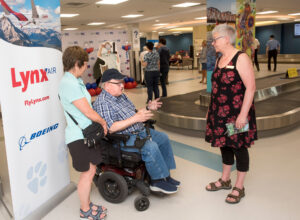 Two people chat with Mayor of New Maryland Judy Wilson-Shee in front of a Lynx Air poster at the Fredericton International Airport
