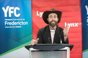 Ryan (Crash) Barton speaks at a podium in front of Lynx and Fredericton International Airport banners