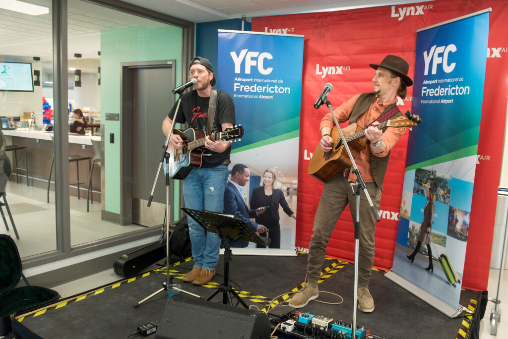 Two musicians from the band Acoustic Ease perform with guitars in front of banners with the Lynx Air and YFC Fredericton International Airport logos