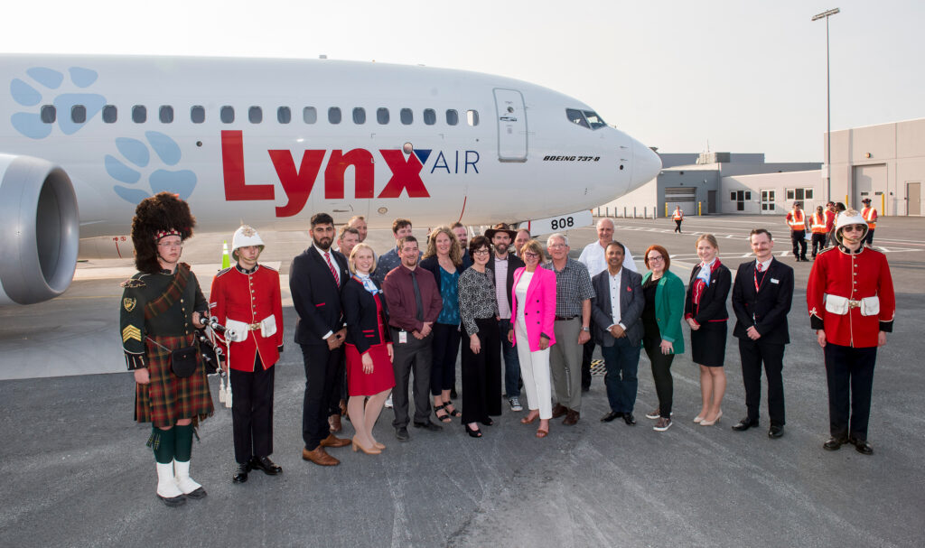 A crowd of people including employees of the Fredericton International Airport Authority and Lynx Air, two guards and a bagpiper from the City of Fredericton, stand in front of a Lynx Air plane.