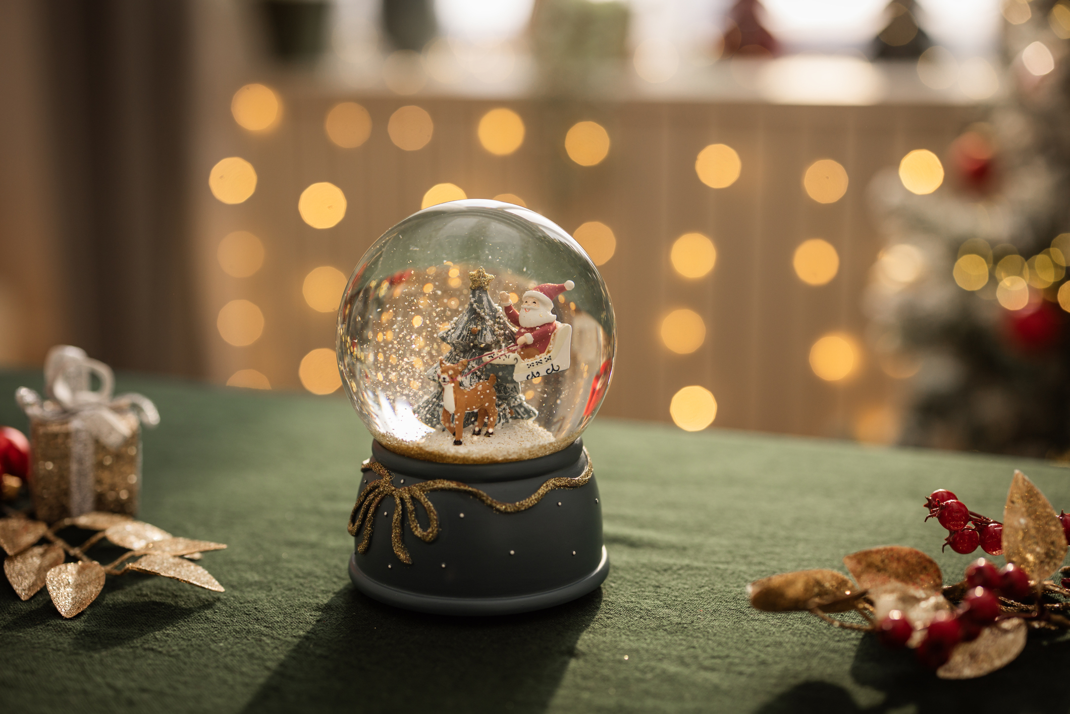 Une boule à neige est posée sur une table verte avec des lumières en arrière-plan. Elle contient un arbre vert avec une étoile au sommet, un père Noël et un renne.